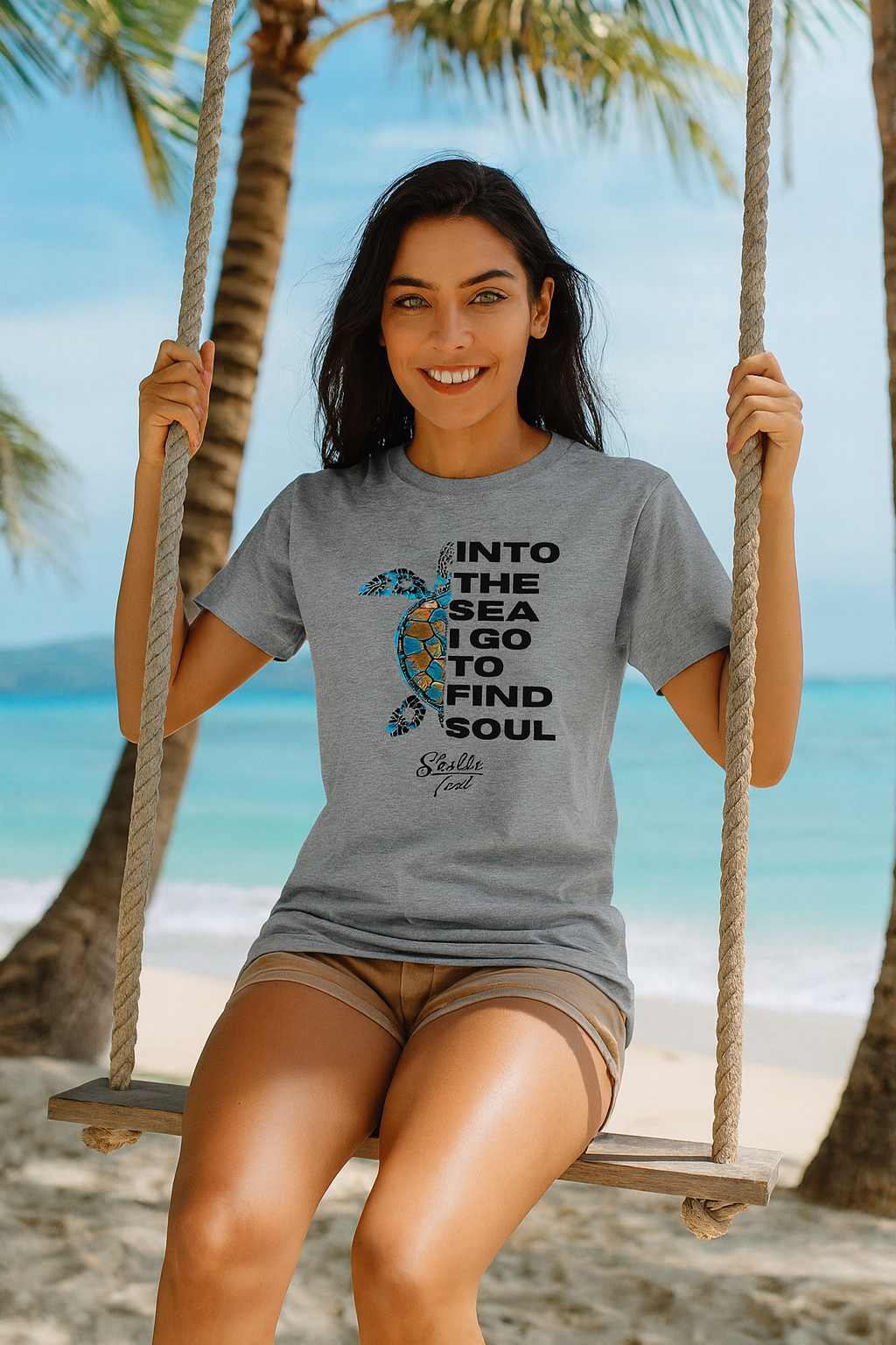 Woman on a swing at the beach wearing a gray t-shirt with a graphic and text.